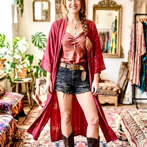 Könnte beinhalten: Eine Frau trägt einen roten Kimono-Stil-Robe über einem Satin-Top und Jeansshorts. Sie trägt auch einen braunen Gürtel, Cowboystiefel und türkisfarbenen Schmuck. Der Raum hat eine böhmische Ästhetik mit gemusterten Teppichen und Vintage-Spiegeln.