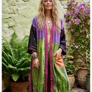 Könnte beinhalten: Ein farbenfrohes, langes, offen geschnittenes Kimono-Kleid im Patchwork-Design. Die Robe hat Bahnen in Schwarz, Lila, Grün und Orange. Das Model trägt mehrere Halsketten und Armbänder. Der Hintergrund zeigt Pflanzen und eine Steinmauer.
