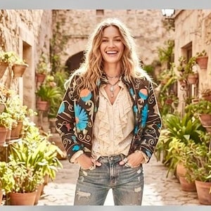 May include: A black jacket featuring vibrant blue and orange floral embroidery, worn over a cream-colored ruffled blouse with a button-down front, paired with light-wash blue denim jeans that have a distressed finish and visible silver rings.