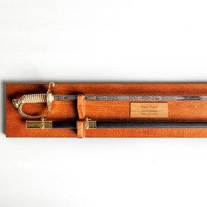 May include: A wooden wall mount display for a sword with a brass plaque for engraving. The plaque has the text "Your Name", "Service Branch", and "Years of Service". The display also features a small American flag.