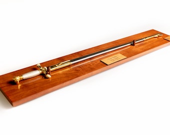 May include: A ceremonial sword with a silver-toned grip and gold-toned crossguard, pommel, and scabbard, mounted on a rectangular wooden display plaque. A small brass plate on the wood features the engraved text "Last Name" and "Date of Award".