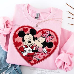 Op de afbeelding: Een lichtroze sweatshirt met een hartvormig ontwerp met Mickey en Minnie Mouse die elkaar omhelzen. Het ontwerp is rood omlijnd met rode harten. Het sweatshirt heeft het merklabel "Gildan".