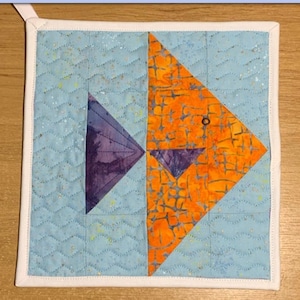 Puede incluir: Un cojín térmico acolchado azul y blanco con un diseño de pez ángel naranja y morado. El cojín térmico es cuadrado de 8,5 pulgadas y tiene un borde blanco. El texto "Angel Fish Heat Pad 8.5" square" está en la parte superior de la imagen. El texto "HOMELY HANDMADEs" está en la parte inferior de la imagen.