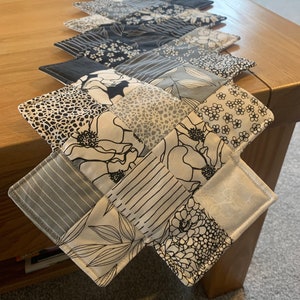 May include: A black and white table runner made of fabric squares with different patterns, including floral, geometric, and abstract designs. The runner is laid out on a wooden table.