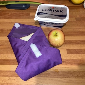May include: A purple reusable food wrap with a white velcro closure, a wrapped sandwich, and a red apple on a wooden surface. A tub of Lurpak spreadable butter is in the background.