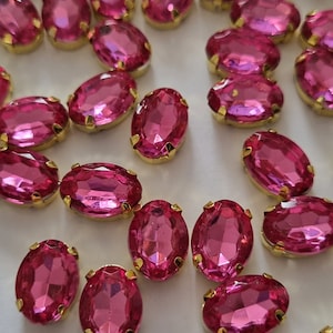 May include: A collection of pink oval-shaped rhinestones with gold settings. The rhinestones are scattered on a white surface.