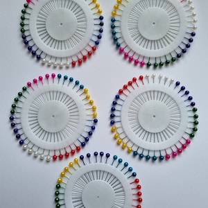 May include: Five circular pincushions with colorful pins. Each pincushion has a white base with a circle in the center and a ring of pins around the edge. The pins are in different colors, including red, blue, green, yellow, and pink.