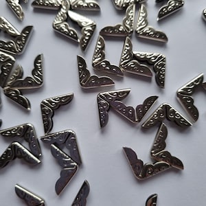 May include: A collection of silver metal corner protectors with a decorative design. The corner protectors are scattered on a white surface.