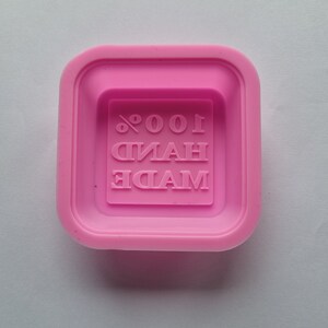 May include: Pink silicone mould for making soap. The mould has a square shape and features the text "MADE 100% HAND" embossed inside.