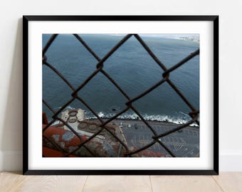 May include: A framed photograph featuring a view of the ocean through a chain-link fence. The water is a deep blue, and a parking lot and structures are visible below. The fence is rusty, and the frame is black.