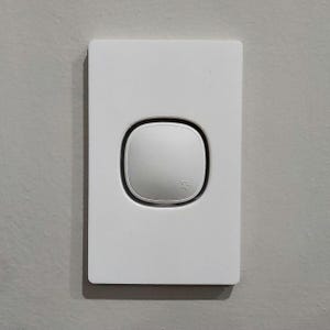 Puede incluir: Interruptor de luz blanco montado en la pared con un botón redondo. El botón tiene un pequeño símbolo que parece una señal de Wi-Fi.
