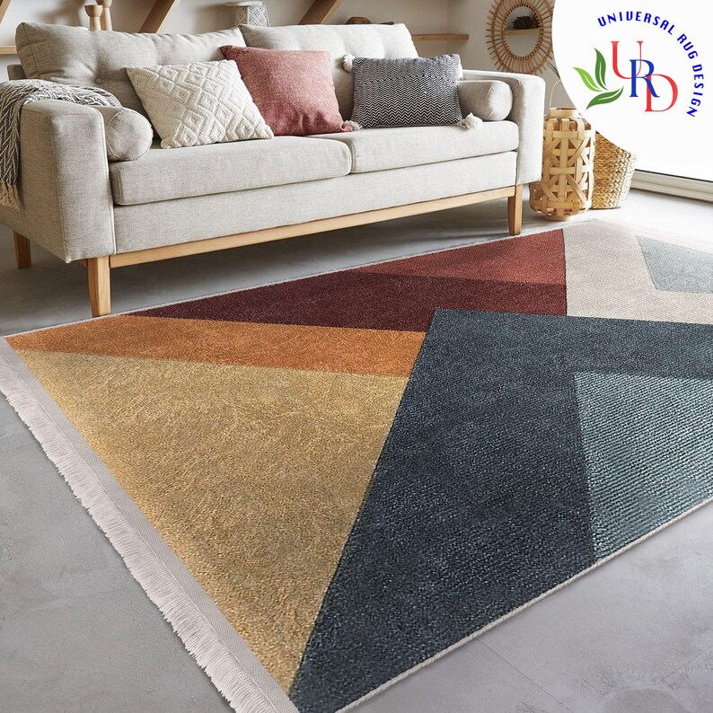 May include: Geometric patterned area rug with a mix of colors including gold, rust, and navy blue. The rug has a fringed edge and is placed on a gray floor. The rug is in front of a beige sofa with decorative pillows.