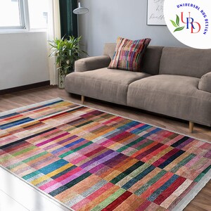 May include: A colorful rectangular rug with a geometric pattern of horizontal stripes in various shades of red, pink, blue, green, and brown. The rug is laid out on a wooden floor in a living room setting with a sofa in the background.