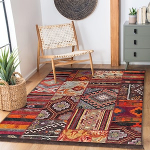 May include: A rectangular patchwork rug with a variety of geometric and tribal patterns in shades of red, orange, brown, and blue. The rug is placed on a wooden floor, with a woven chair and a green cabinet in the background.