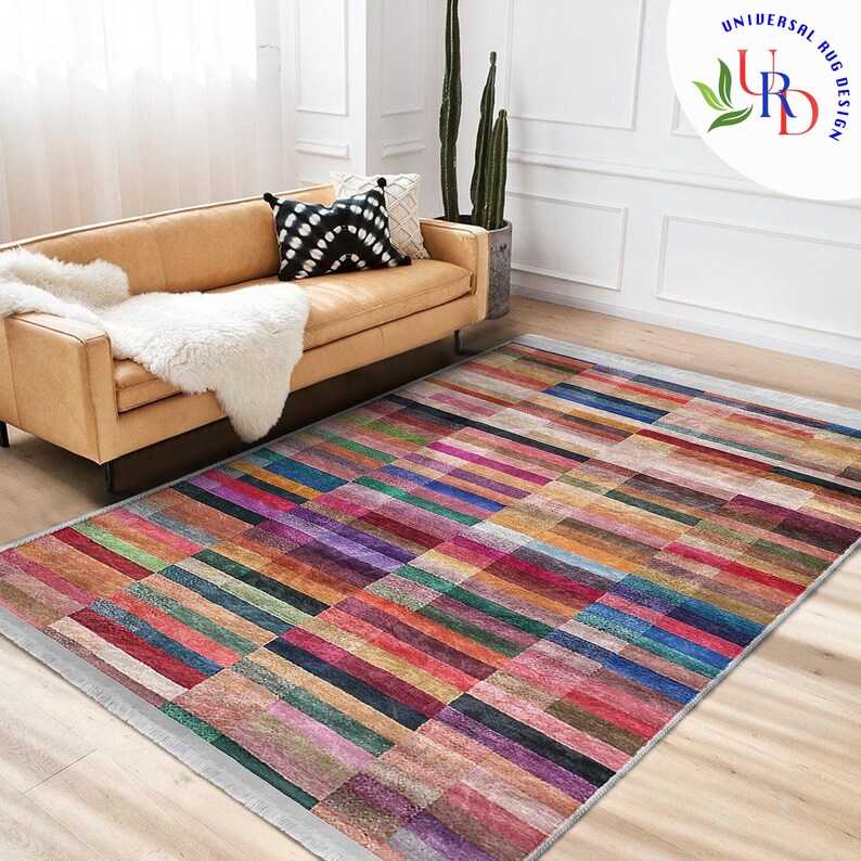 May include: A colorful rectangular rug with a geometric pattern of horizontal stripes in various shades of red, pink, purple, blue, green, and brown. The rug has a white border and is laid out on a light brown wooden floor.
