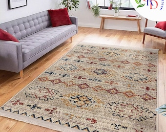 May include: A rectangular area rug with a beige background and a geometric pattern in red, blue, and gold. The rug has a fringe on the short ends and is placed on a hardwood floor. The rug is in a living room setting.