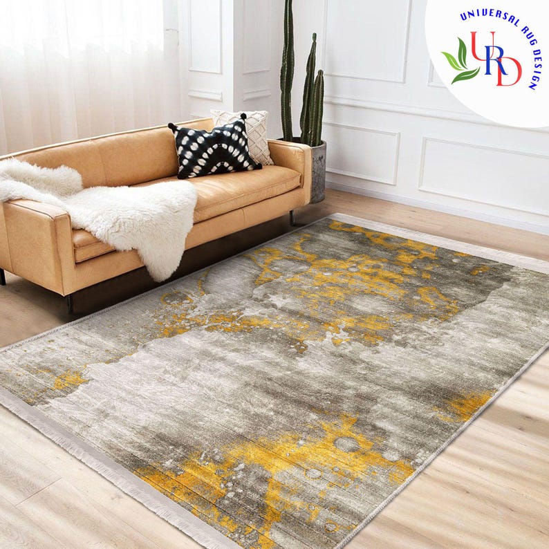 May include: A rectangular area rug with a gray and yellow abstract design. The rug features a blend of gray tones with splashes of yellow, creating a modern aesthetic. The rug is placed on a wooden floor, near a tan sofa.