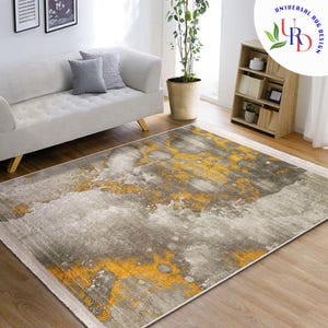 May include: A rectangular area rug with a gray and yellow abstract design. The rug has a distressed, mottled appearance with splashes of yellow against a gray background. The rug is placed on a hardwood floor in a living room setting.