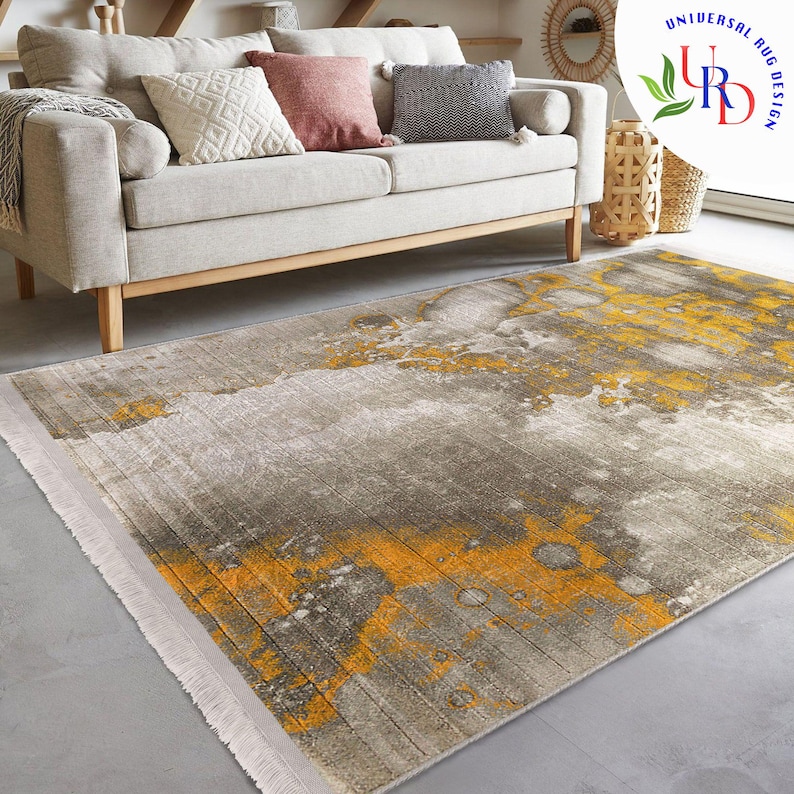 May include: A rectangular area rug with a distressed design in shades of gray and yellow. The rug has a fringe on the short ends and is placed on a gray floor. A beige sofa with pillows is in the background.