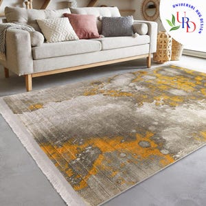 May include: A rectangular area rug with a distressed design. The rug features a gray and yellow color palette with a textured appearance. The rug is placed on a gray floor in front of a beige sofa with decorative pillows.