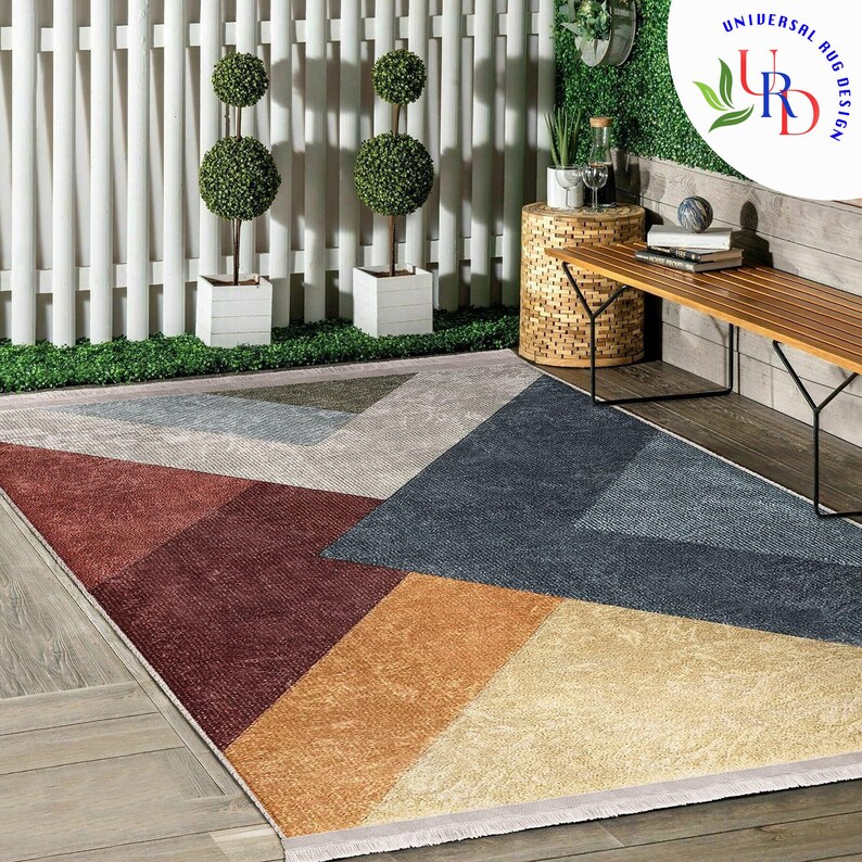 May include: A geometric patterned area rug with a mix of colors including red, blue, tan, and gray. The rug is rectangular and has a modern design. The rug is placed on a wooden deck, with a white fence and greenery in the background. The rug is from Universal Rug Design.