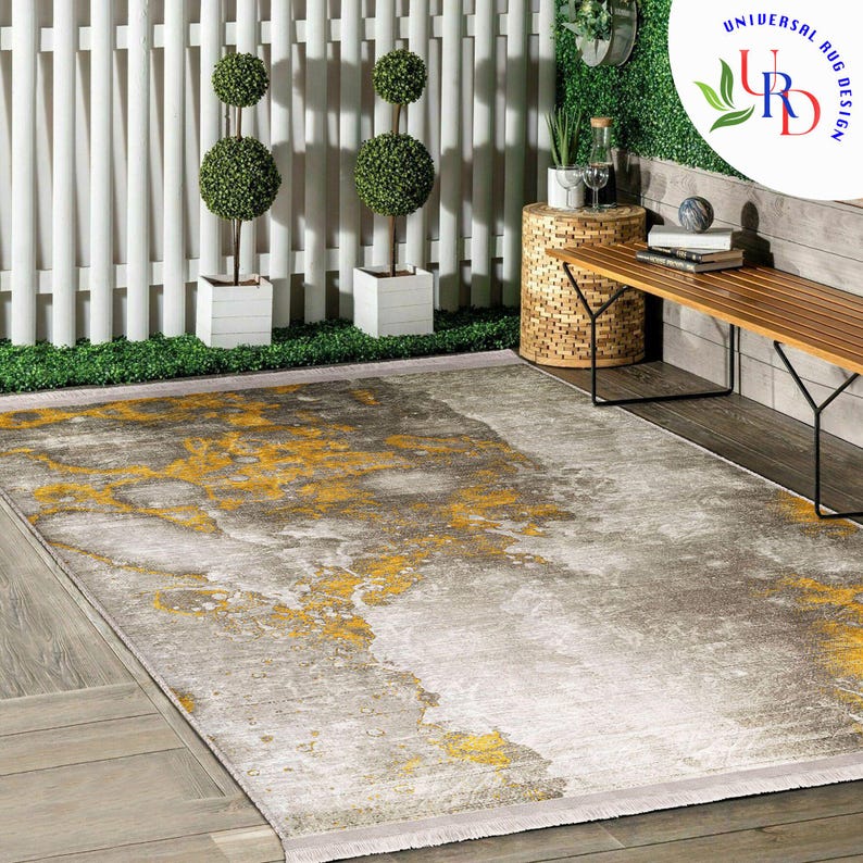 May include: A rectangular area rug with a gray and yellow abstract design. The rug has a light fringe and is placed on a wooden deck. The background includes a white fence, topiary trees, and a bench.