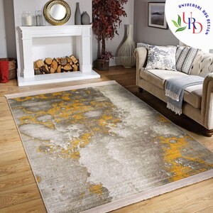 May include: A rectangular area rug with a modern design. The rug features a gray and yellow abstract pattern. The rug is placed on a hardwood floor in a living room setting. The rug has a fringe border and is under a sofa.
