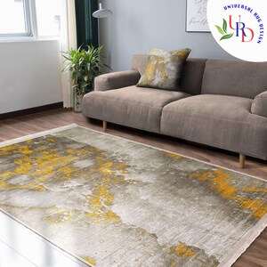 May include: A rectangular area rug with a gray and yellow abstract design. The rug is on a hardwood floor in front of a gray sofa and a decorative pillow. The rug has a fringe on the short ends.