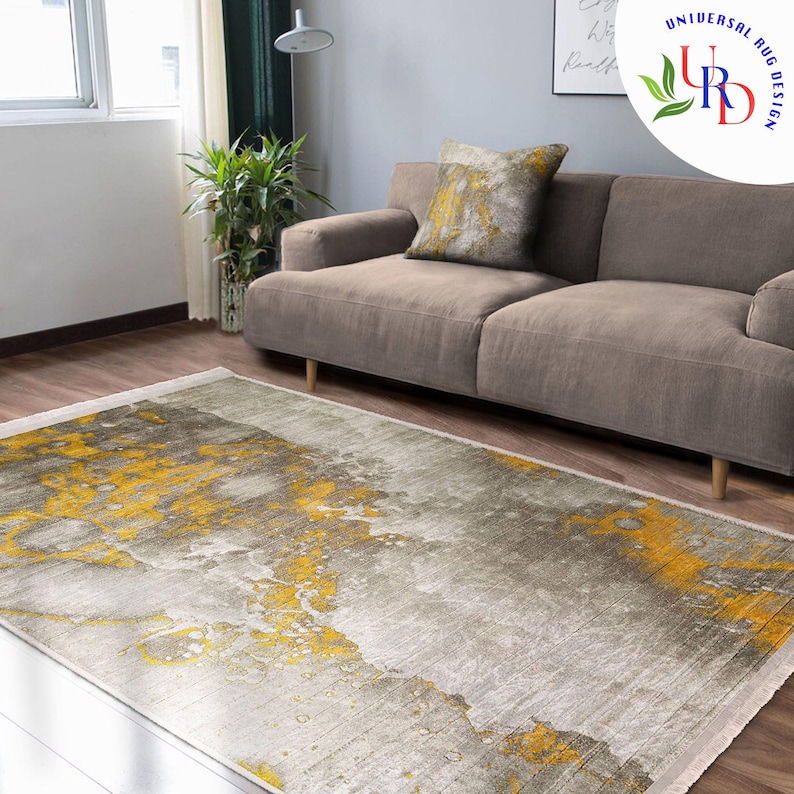 May include: A rectangular area rug with a gray and yellow abstract design. The rug is on a hardwood floor in front of a gray sofa and a decorative pillow. The rug has a fringe on the short ends.