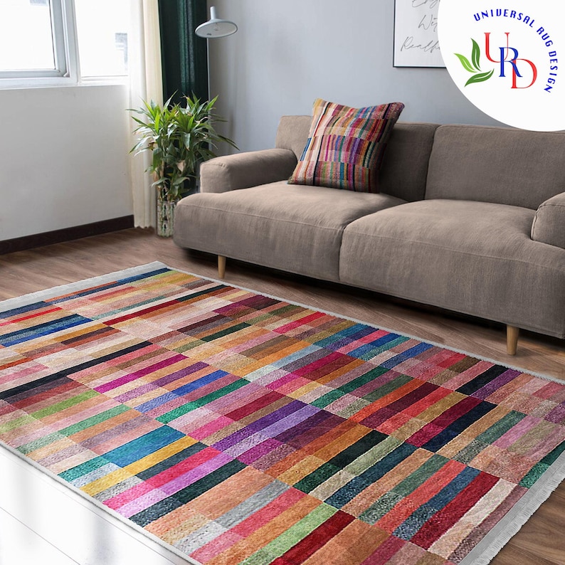 May include: A colorful rectangular rug with a geometric pattern of horizontal stripes in various shades of red, pink, blue, green, and brown. The rug is laid out on a wooden floor in a living room setting with a sofa in the background.