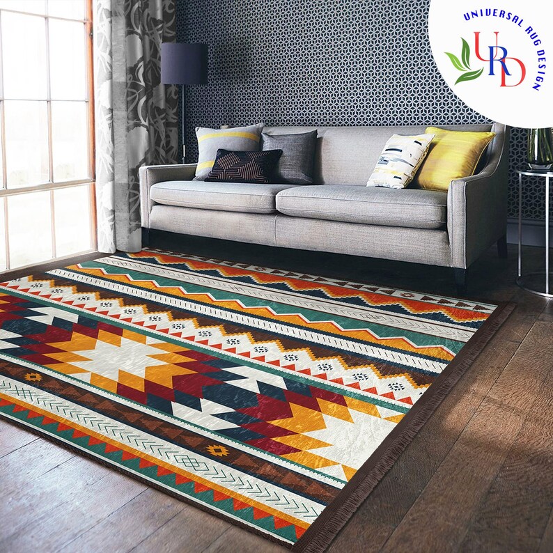 May include: A colorful geometric patterned rug with a brown fringe. The rug is laid out on a hardwood floor in front of a gray sofa with pillows.