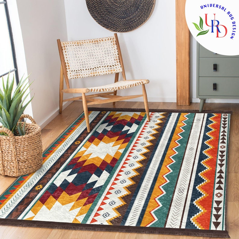 May include: A white and brown area rug with a geometric pattern in shades of red, yellow, blue, and green. The rug has a fringe border and is laid out on a wooden floor with a chair in the background.