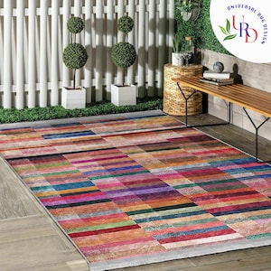 May include: A colorful rectangular area rug with a geometric pattern of multicolored stripes. The rug is laid out on a wooden floor in a patio setting with a wooden bench and potted plants.