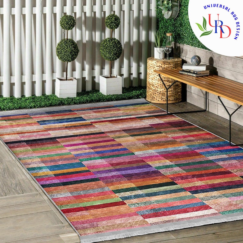 May include: A colorful rectangular area rug with a geometric pattern of multicolored stripes. The rug is laid out on a wooden floor in a patio setting with a wooden bench and potted plants.