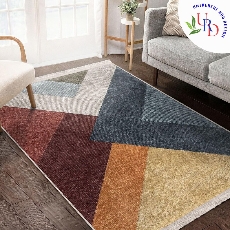 May include: Geometric patterned area rug with a mix of colors including red, blue, orange, and beige. The rug has a rectangular shape and is placed on a hardwood floor. The design features angular shapes and a white fringe.