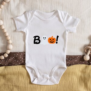 Pode incluir: Macac&atilde;o branco para beb&ecirc; com um gr&aacute;fico preto de "BOO!" que apresenta um fantasma e uma ab&oacute;bora.