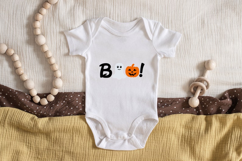 Pode incluir: Macac&atilde;o branco para beb&ecirc; com um gr&aacute;fico preto de "BOO!" que apresenta um fantasma e uma ab&oacute;bora.