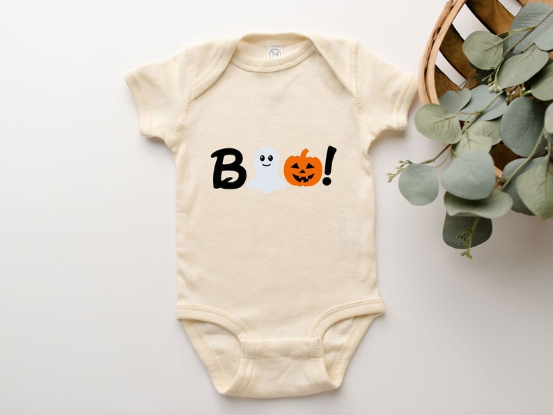 Pode incluir: Um macac&atilde;o de beb&ecirc; cor creme com um gr&aacute;fico preto de "BOO!" que apresenta um fantasma e uma ab&oacute;bora.