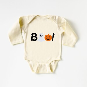 Pode incluir: Um macac&atilde;o de beb&ecirc; de manga comprida de cor creme com um gr&aacute;fico preto, branco e laranja que diz "BOO!" com um fantasma e uma ab&oacute;bora.