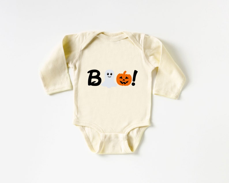 Pode incluir: Um macac&atilde;o de beb&ecirc; de manga comprida de cor creme com um gr&aacute;fico preto, branco e laranja que diz "BOO!" com um fantasma e uma ab&oacute;bora.