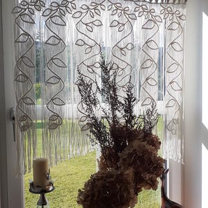 May include: A macrame window treatment with a leaf design hangs in front of a window. A candle holder, a vase of dried flowers, and a wooden deer figurine sit on the windowsill. The window looks out onto a green lawn.