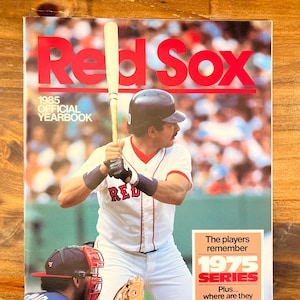 May include: The cover of the 1985 Red Sox Official Yearbook. The cover features a baseball player in a white uniform with red accents, holding a bat. The text "Red Sox" is in large red letters. A sticker reads "1975 Series".