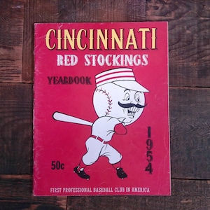 May include: A red and white vintage 1954 Cincinnati Red Stockings yearbook featuring a cartoon mascot with a baseball for a head. The text "First Professional Baseball Club in America" is at the bottom of the cover.