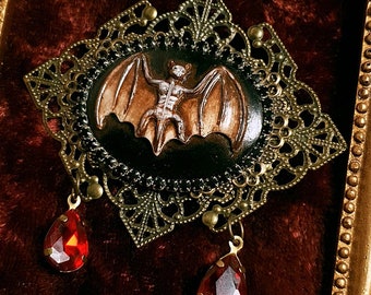 May include: A gold-toned ornate frame with a black and brown bat brooch with red gemstone accents. The brooch is in the shape of a bat with its wings spread out.