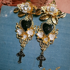 May include: Gold tone earrings with black heart charms, white pearl-like flowers, and black cross accents.