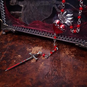 May include: A silver-toned dagger with a red stripe and a red beaded chain. A red heart pendant with silver wings and a red beaded chain are also visible. The items are displayed on a textured, brown surface.