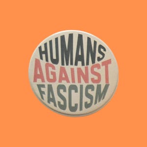 May include: A circular pinback button with a cream-coloured background. The text HUMANS is printed in dark grey at the top, AGAINST in red in the centre, and FASCISM in dark grey at the bottom, all in a bold, sans-serif, capitalised font.