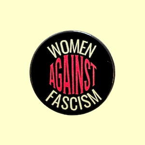 May include: A circular black badge with the text "WOMEN AGAINST FASCISM" in white and red. The words are arranged in a circular design, with "WOMEN" at the top, "AGAINST" in the middle, and "FASCISM" at the bottom.
