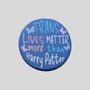 May include: A circular blue button featuring the text "TRANS LIVES MATTER more than Harry Potter" in a mix of white, light blue, and light pink lettering. The design is decorated with four small butterflies in shades of light blue and light pink.
