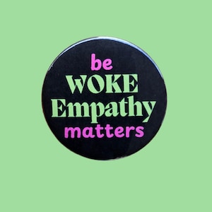 May include: Black button with the text "be woke empathy matters" in pink and green lettering.
